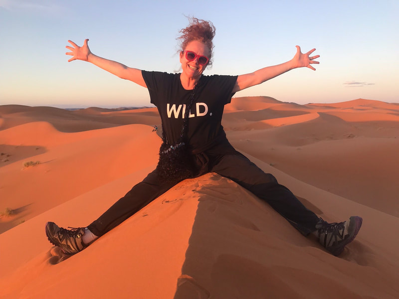 Jill Gleeson, smiling with her arms spread wide, on top of a sand dune in the Sahara, Morocco